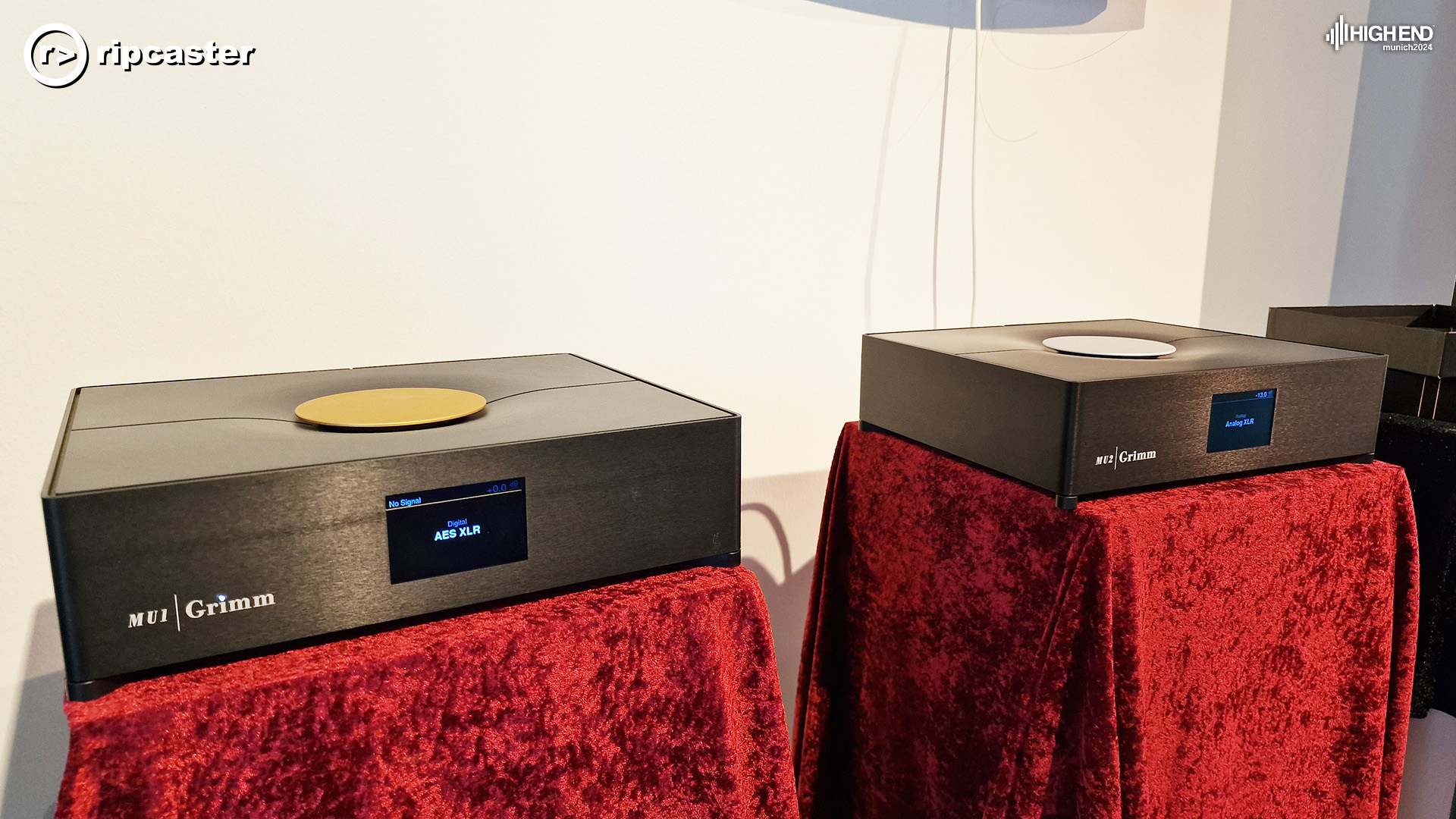 Two Grimm audio units on plinths with red velvet draped over.  The back wall is cream.  The unit to the left is an MU1.