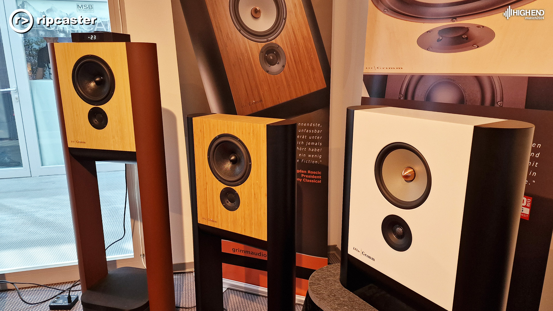 Three Grimm speakers.  Two on standis and the white on closest to us is on a plinth.  There are large pictures of speakers behind these real speakers