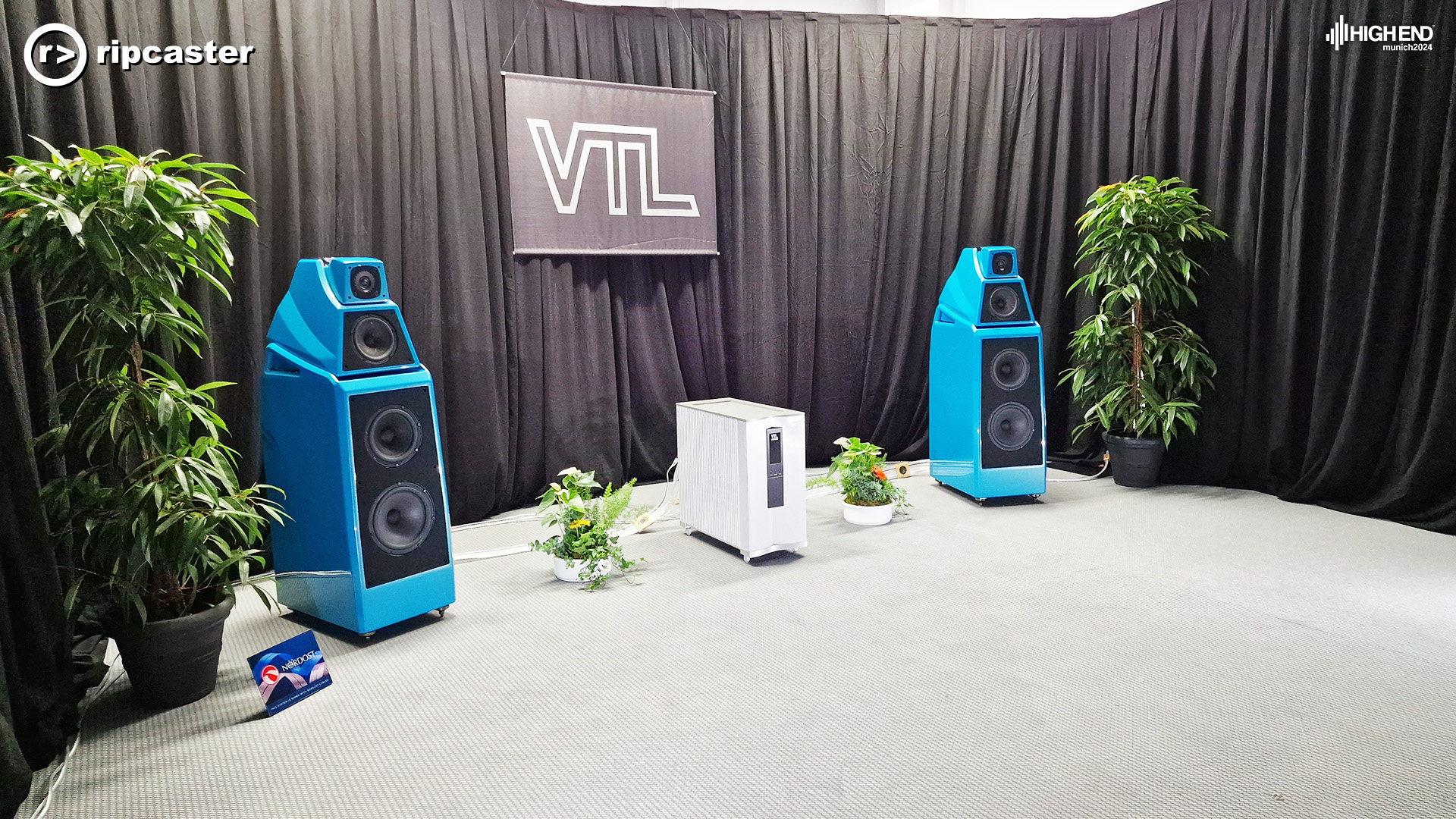 VTL blue floorstanding speakers with black curtains behind and tall plants either side of the speakers