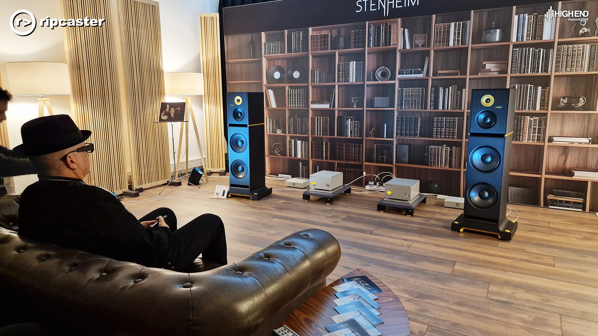 A man in the foreground sitting down and a pair of floorstanding speakers in front of him either side of two low HiFi boxes