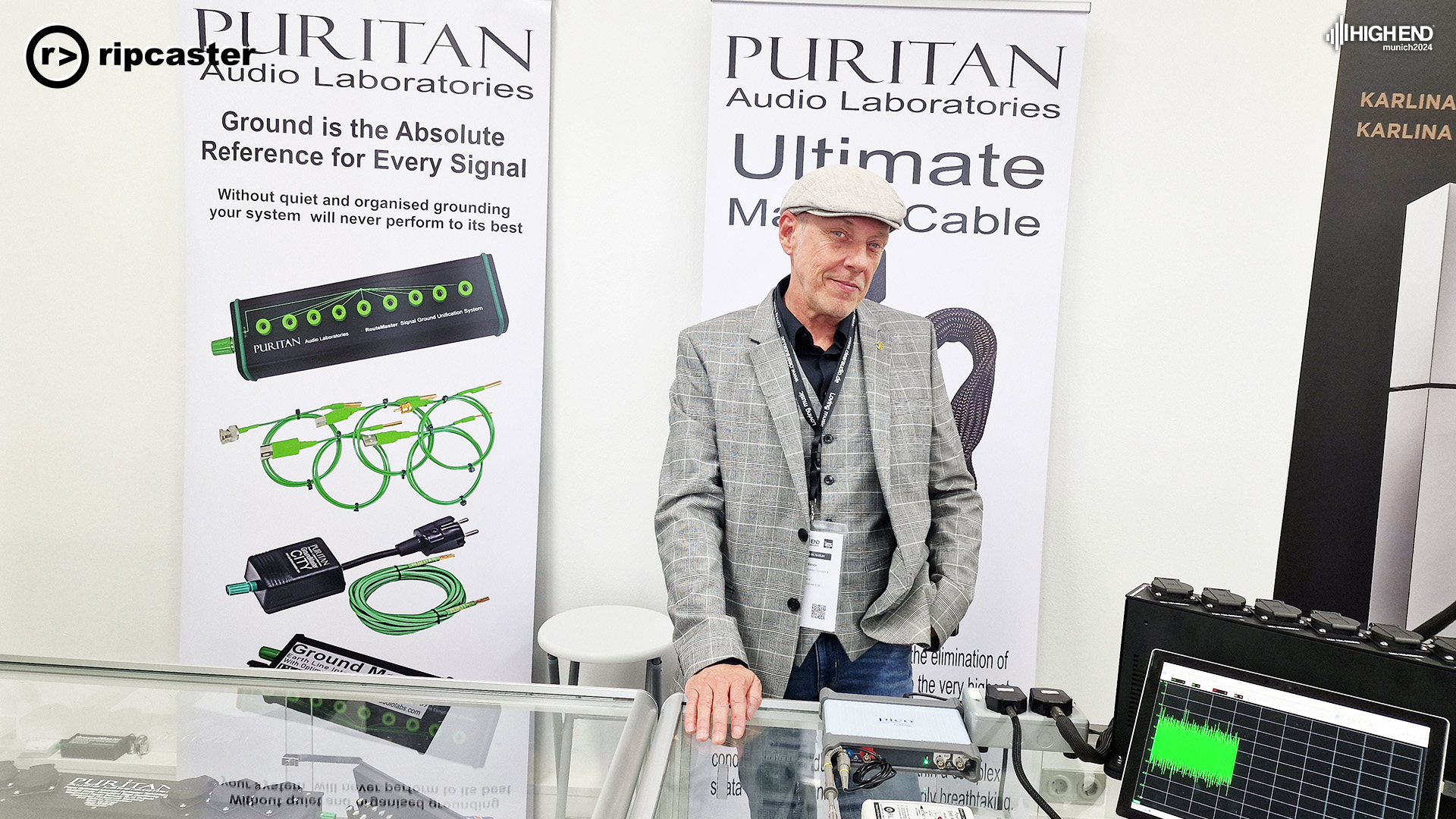 A very well dressed guy in a flat cap, waistcoat and jacket standing behind a glass cabinet at the Puritan stand.