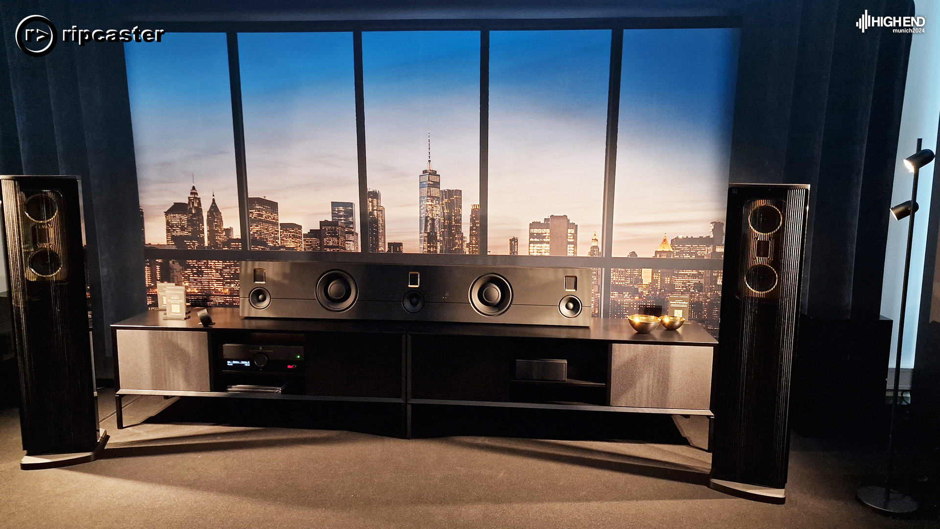 A pair of black floorstanding speakers in front of a large city backdrop with HiFi equipment between