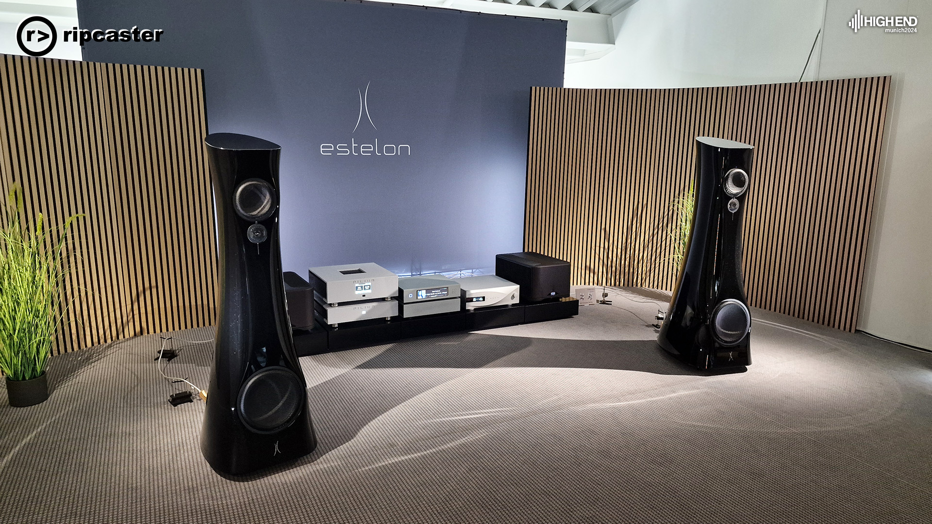 A pair of black floorstanding speakers.  Estelon.  Various pieces of HiFi kit between the speakers on a low stand.  there's acoustic panels either side of the room.