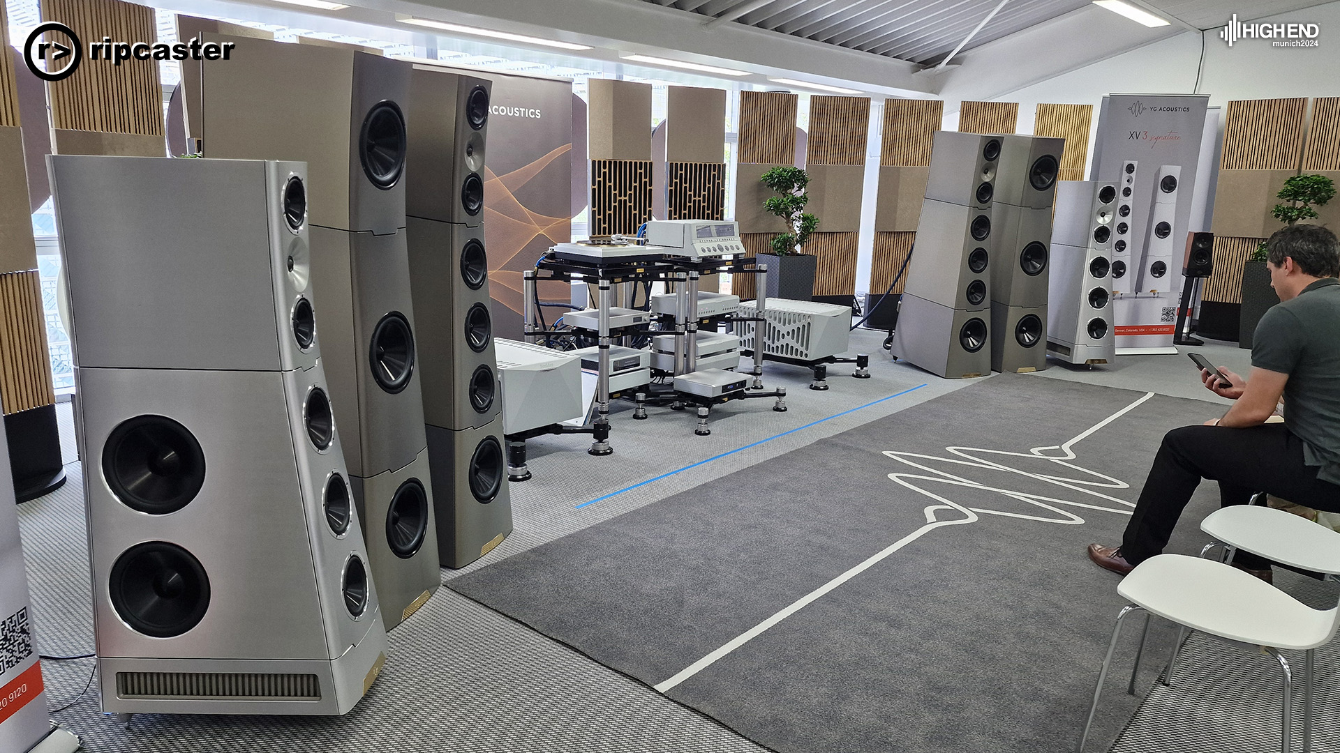 A lot of floorstanding speakers in silver with lots of other HiFi equipment and a man sitting on a seat to the right.