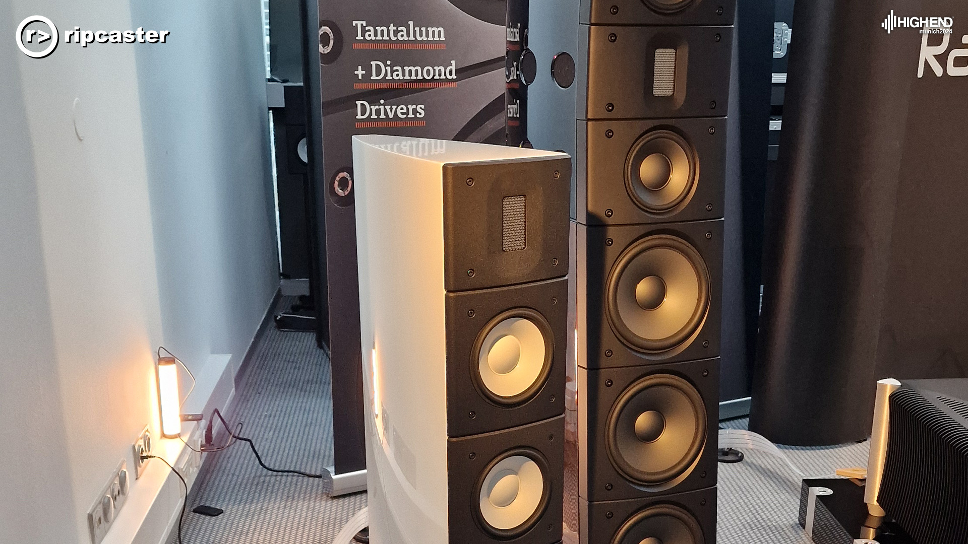Two Raidho speakers beside each other.  One is taller than a person, the other is smaller and white.  There are Raidho signs in the background
