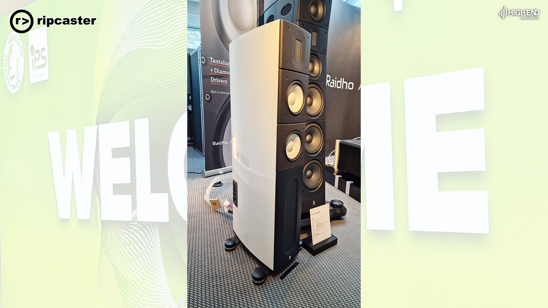 This is the new Raidho X2.6 speaker in white beside a much larger speaker on its right.  Both are floorstanding.  There are signs in the background.
