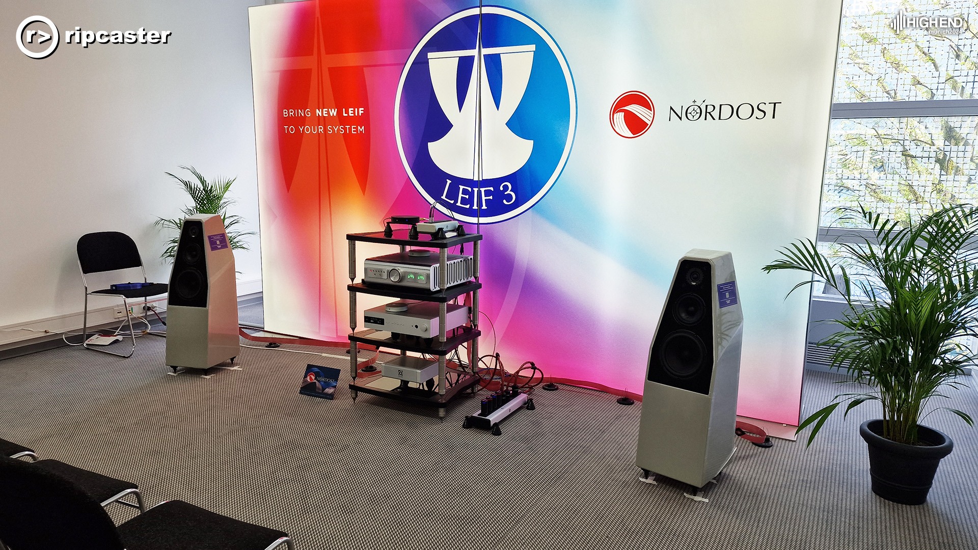 A big Nordost backdrop with a pair of speakers either side of a stack of HiFi equipment.  There are chairs visible in the foreground on the left
