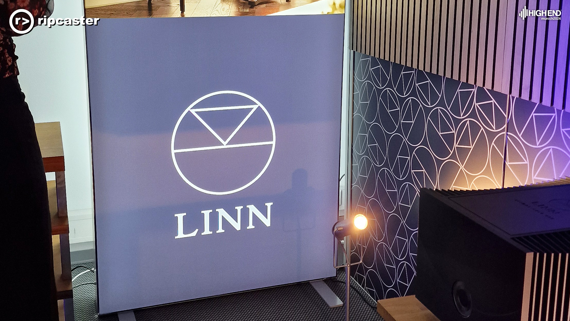 The Linn logo on a purple background with lot of Linn logos on a sign beside it
