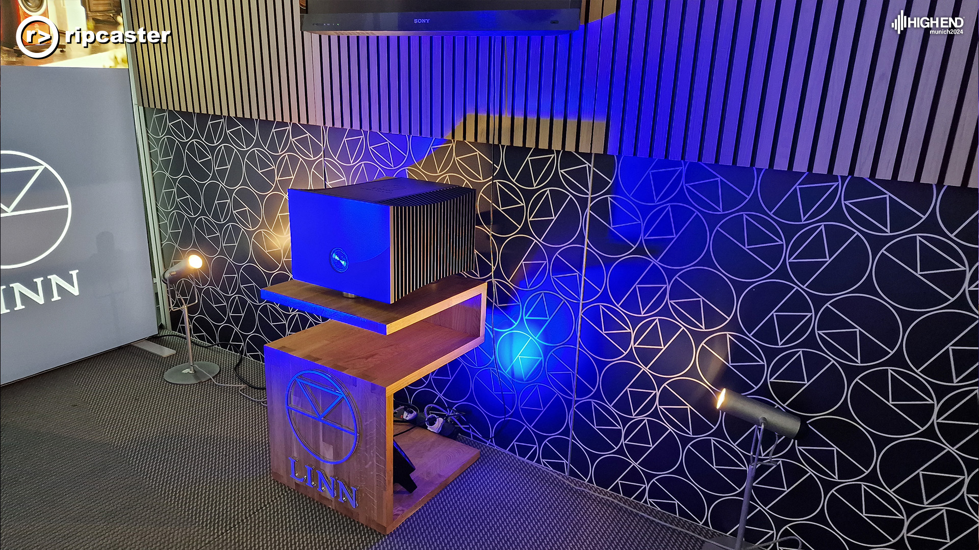 A Linn Klimax Solo 800 on a wooden stand in front of a backdrop of many Linn logos and a Linn sign to the left.  The Klimax Solo 800 is a black rectangular box shape that reflects light (in this case a blue)
