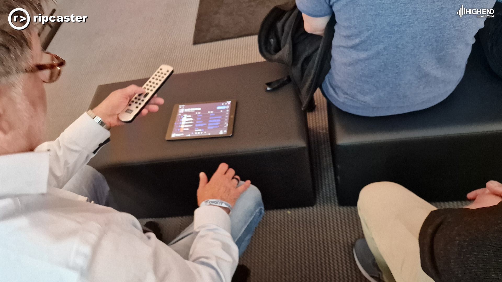 A person holding a remote control with an iPad on a seat in front of him.