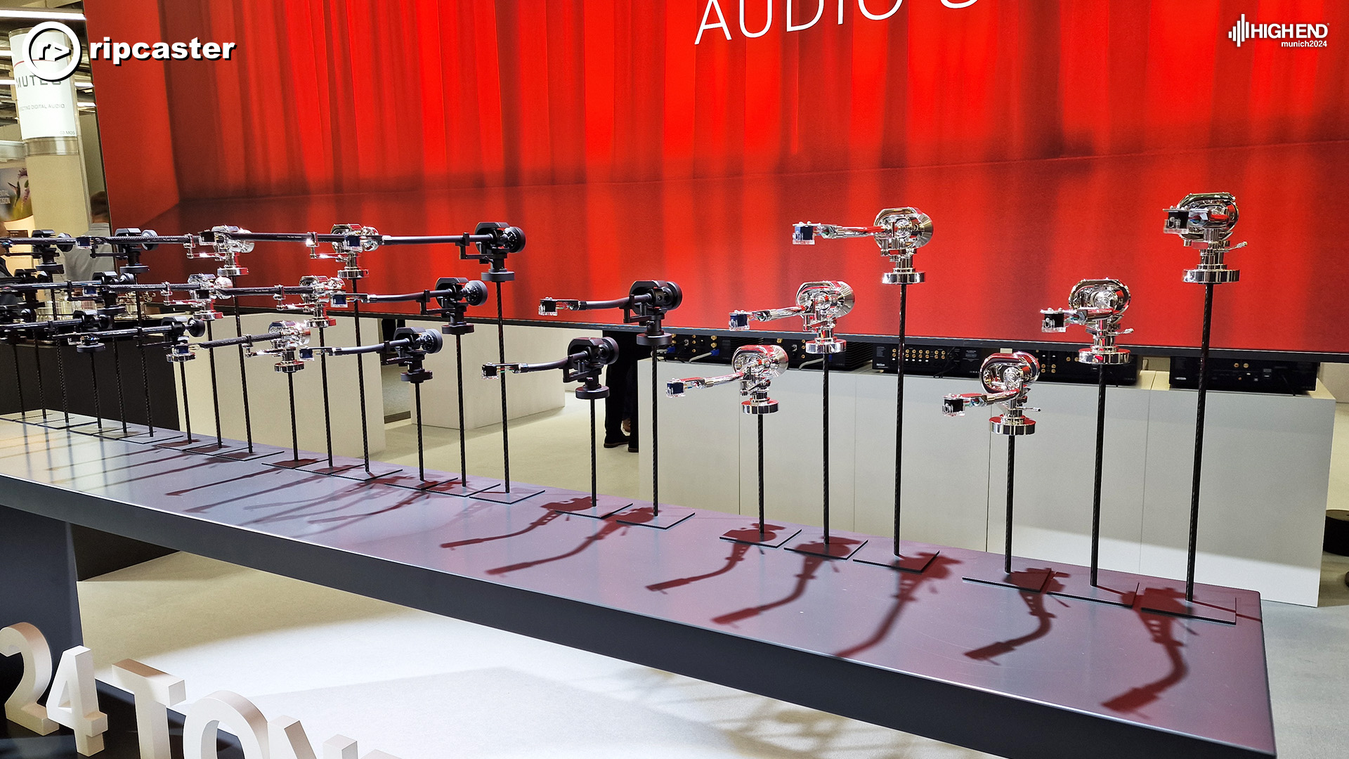 A stunning arrangement of Tonearms from Project - they are all on long sticks and lined up beside each other at different heights.  There's a red backdrop.