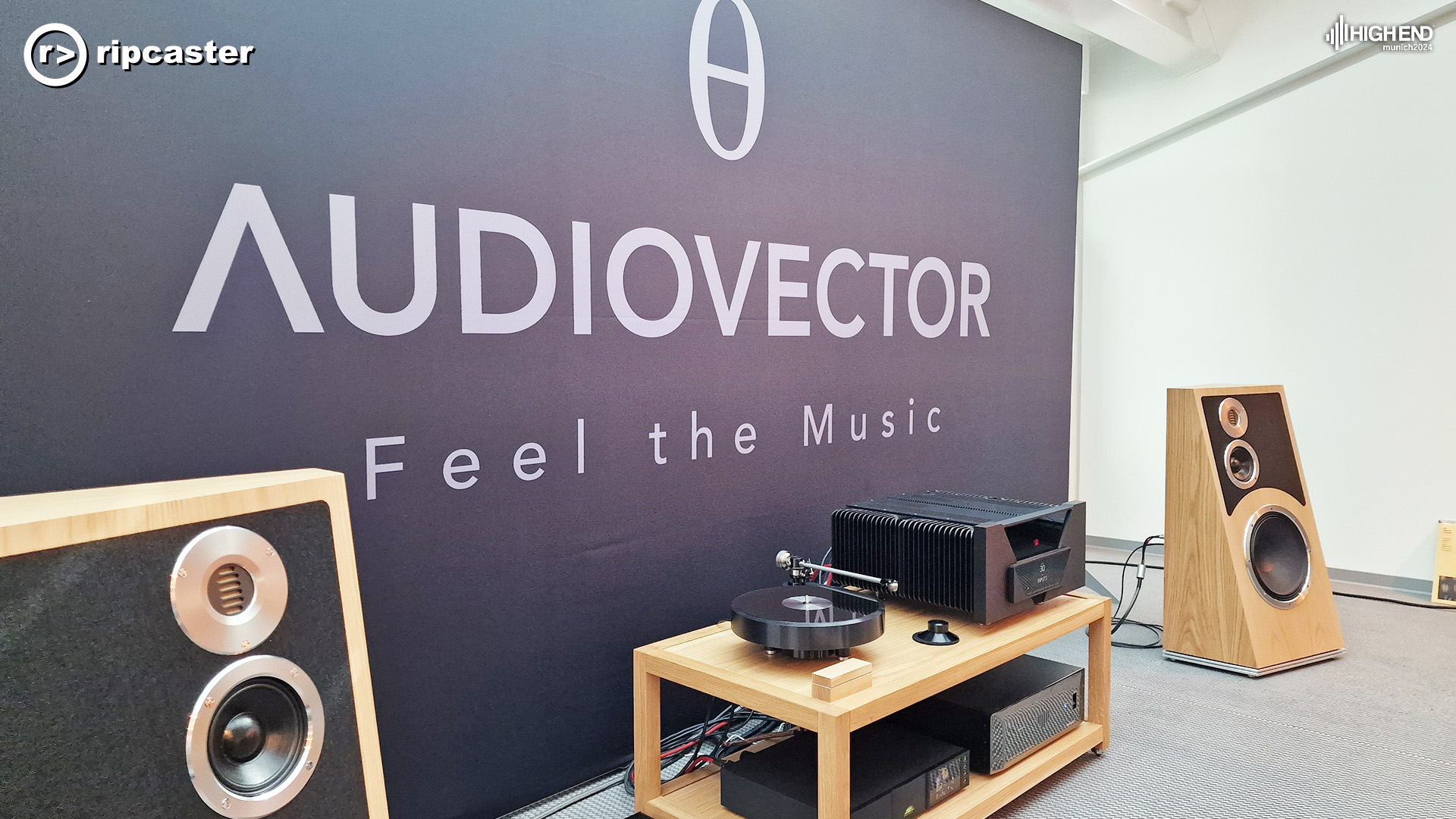Audiovector in big letters on the backdrop with the words "Feel the Music".  A pair of the new Audiovector speakers either side of some HiFi equipment on a low stand.  The new Audiovector speakers have the shape of a traditional wedge standing up on the larger of the two ends