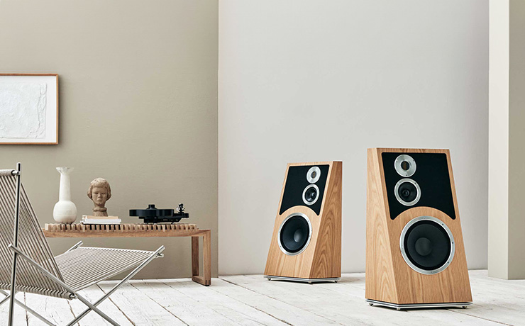 A pair of Audiovector Trapeze speakers in Oak on the right of the image.  On the left is a chair and at the back of the image a low table with a vase, a ceramic head and a turntable on