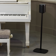 Flexson Floor Stand One/Play1 EU x1 in black with speaker next to a white piano with the piano stool partially visible.  In a room with a taupe tiled floor.