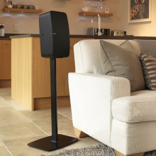 Flexson Floor Stand Play5 x1 in black with Sonos Play:5 next to a sofa with a kitchen area behind.