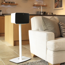 Flexson Floor Stand Play5 x1 in white with a horizontally placed Sonos Play:5 beside a sofa with a kitchen area behind.