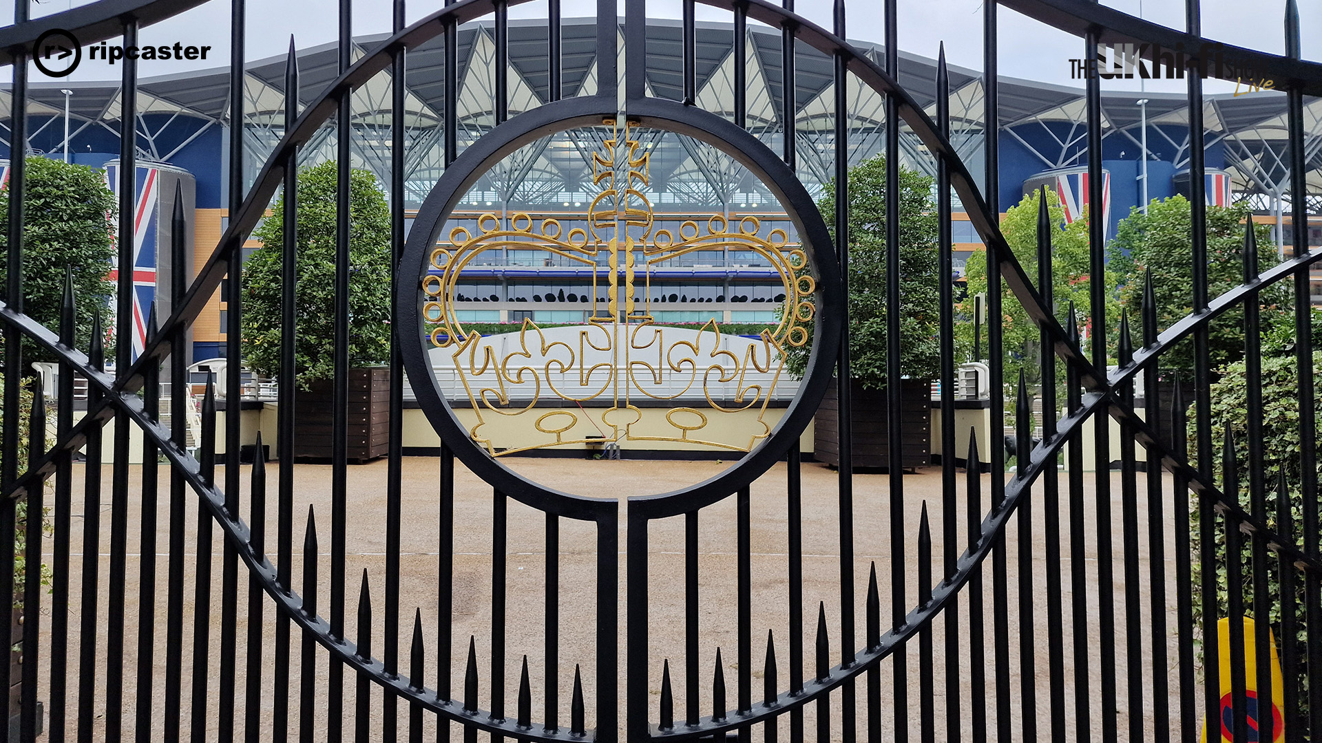 The gate to Ascot.  The gates are black with a gold coloured crown in the centre.  The Ascot building is in the background