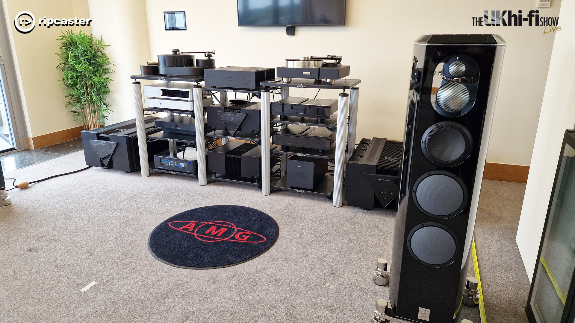 AMG.  A black floorstanding speaker with various pieces of HiFi equipment in the background on various stands.