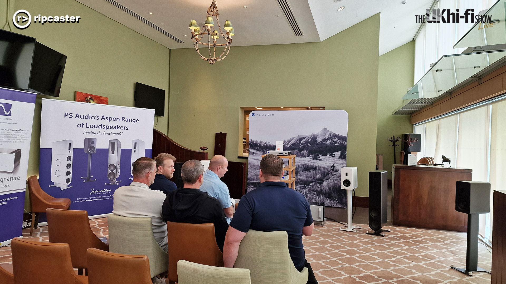 Five men viewed from behind.  In a room with promotional boards for hifi equipment and selected speakers and HiFi equipment