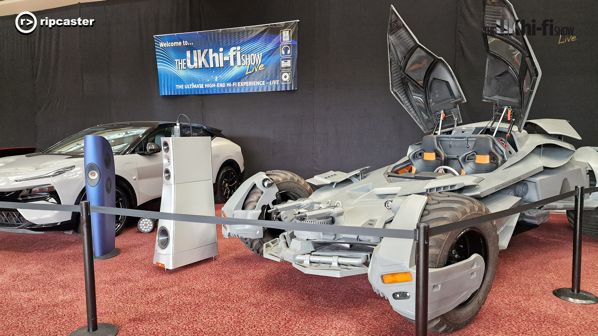 A futuristic looking off-road car with doors that open upwards.  There's a white floorstanding speaker beside it with a white car the other side of the speaker