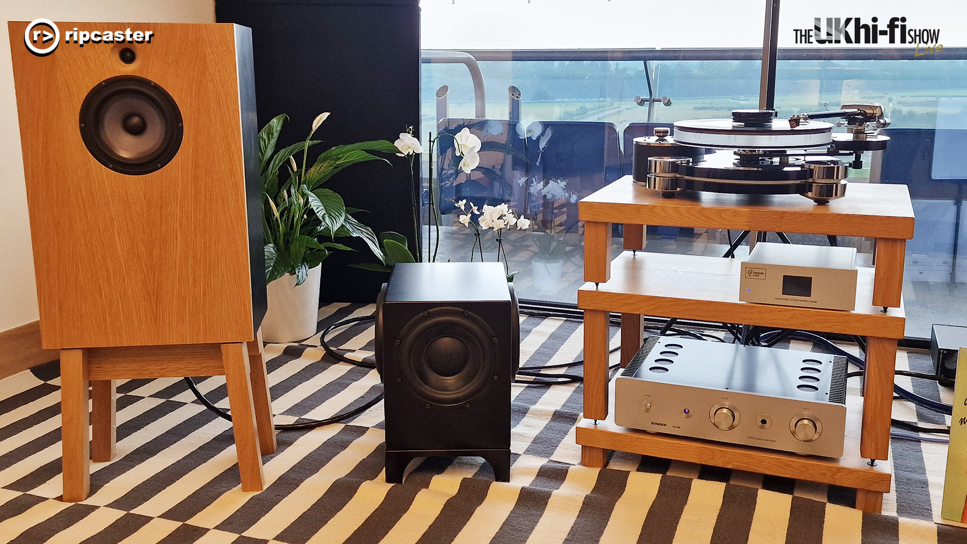 A wooden speaker on wooden legs beside a subwoofer and a rack with a turntable on the top