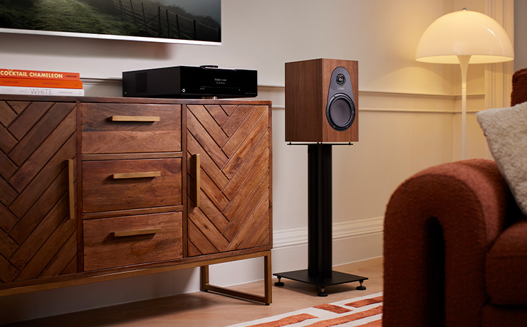 the Linn 119 Loudspeaker in walnut on a stand beside a wooden sideboard with a cream floorstanding mushroom light to the right.