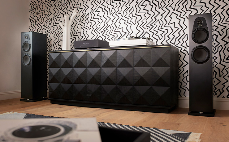 A pair of Linn 150 speakers in satin black either side of a modern sideboard unit.  There are records in the foreground and the sideboard has a turntable and a DSM on top of it along with a white modern looking vase.  The wallpaper is white with lots of black zigzag lines over it