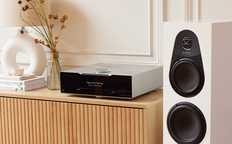 The Linn 150 speaker in white beside a wooden sideboard with a DSM on it, a lamp, books and a vase