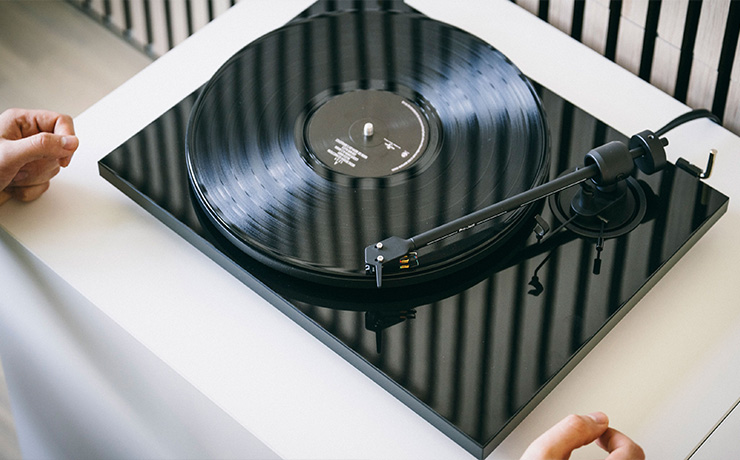 The Project E1.2 BT in a gloss finish with reflections. A vinyl record is playing and the turntable is on a white unit with someone's hands either side of it