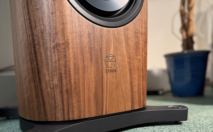 A close-up view of the base of a walnut Linn 360 speaker with a blurred blue plant pot in the background. The Linn logo is nicely in view on the bottom section of the speaker between the bottom driver and the base
