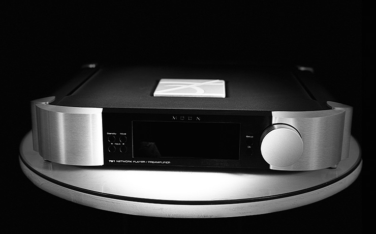 A black and white image of the MOON 791 Network player and preamplifier.  It's standing on a circle of glass that's on top of a white circular table. The light is catching the silver very nicely.
