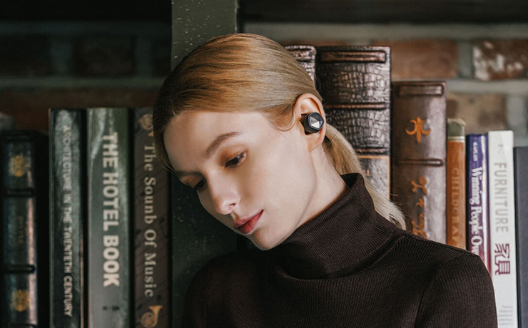 A woman in a black polo-neck top and her hair in a ponytail.  She's in front of a shelf of books and has a UW100MKII earbud in.