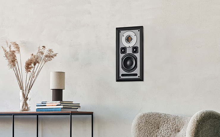 Audiovector Inwall Speaker in black.  To the left of the image is a table with books and a vase on the table.