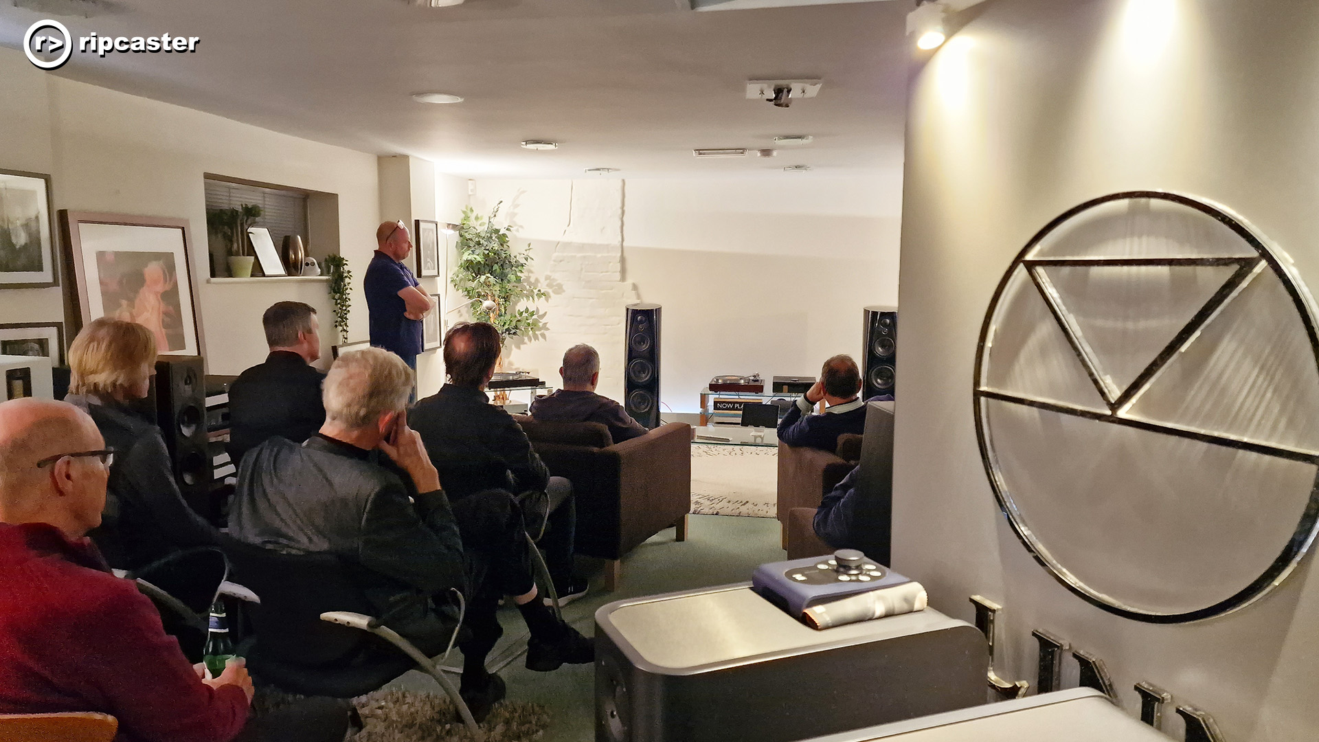 A room full of people viewed from the back.  The people are sitting on chairs in a large room with HiFi equipment.