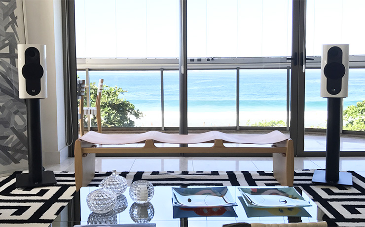 A pair of Kii Three Speakers in white in front of a large window that looks out to a beach with a very clear looking blue sea.  In the foreground there's a coffee table with various glass items on it.  The rug is a geometric black and white pattern and there's a bench with a canvas seat