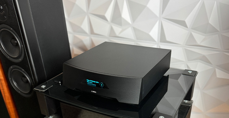 The Lumin P1 Music Player in black with a black speaker to the left of the image and a geometric raised pattern on the back wall behind it.  It's on a glass and black HiFi stand