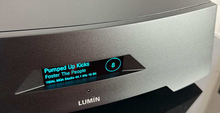 The Lumin P1 Music Player in black although it looks dark silver in this photo.  The display window reads 8 in a circle on the right.  On the left it shows "Pumped Up Kicks", under that "Foster The People" and under that "TIDAL MQA Studio 44.1 kHz 16 Bit