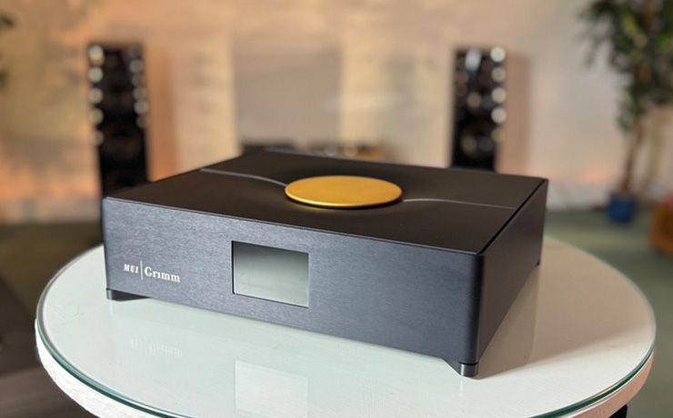 The Grimm MU1 Music Streamer.  It's black with a display window at the front and a gold coloured disc on the top which can be used to control the volume.  There are two floor standing speakers blurred in the background and the MU1 is on a white circular, glass topped, table 