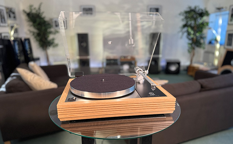 The Linn Majik LP12 MM Turntable with a fluted plinth in oak.  The lid is raised.  Behind the turntable are speakers and sofas along with other HiFi equipment and some large indoor plants