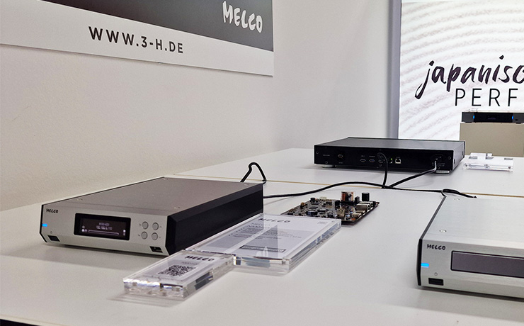 The Melco N100 on a white table at the Munich 2024 HiFi show beside another Melco box on a white table.  The N100 is a silver box that looks the shape of an old answer machine.  It has a screen at the front and four buttons to the right.