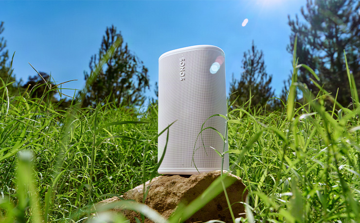 The Sonos Play speaker in white standing on a rock in a field of grass with trees in the distance