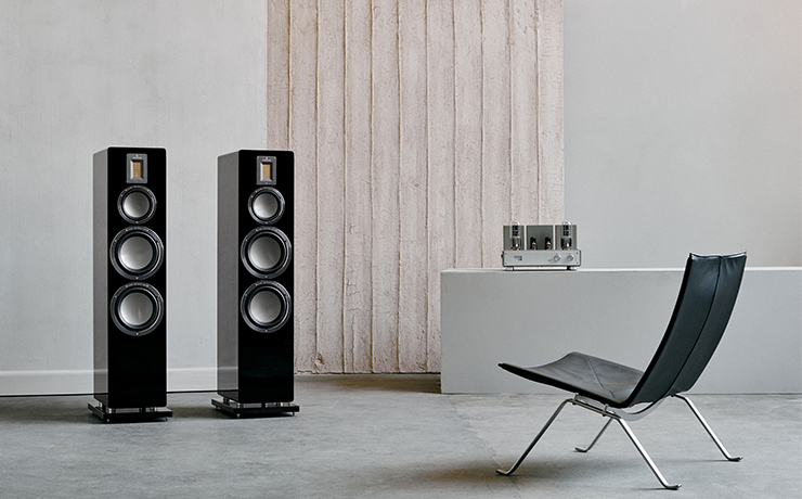 A pair of black QR7 speakers with a leather chair in the room and a valve amplifier