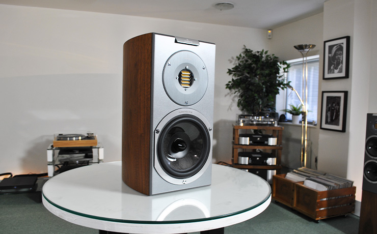 The Audiovector R1 Arrete on a white table in our Checkendon showroom.  There are other pieces of audio equipment in the background and some vinyl records in an orange crate style container.  Black and white photos are on the wall.