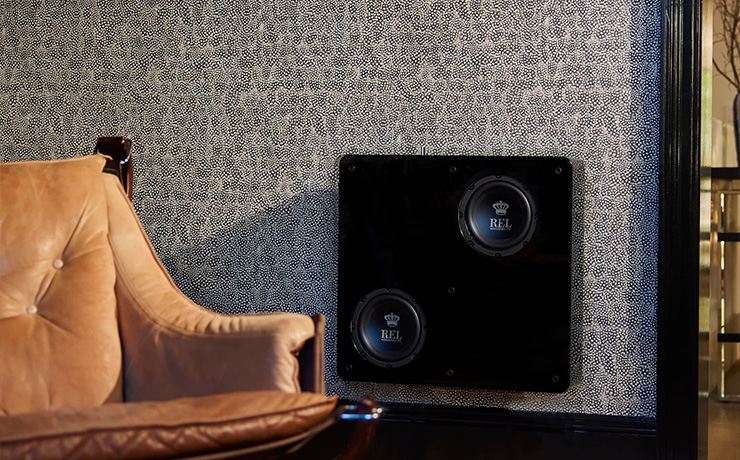 The REL PL1 in black mounted on a wall in a living space.  there is a leather chair to the left of it.