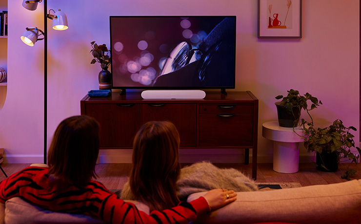 Sonos Ray on a tv stand in front of a tv.  There are two people in the foreground with their backs to the camera sitting on a sofa