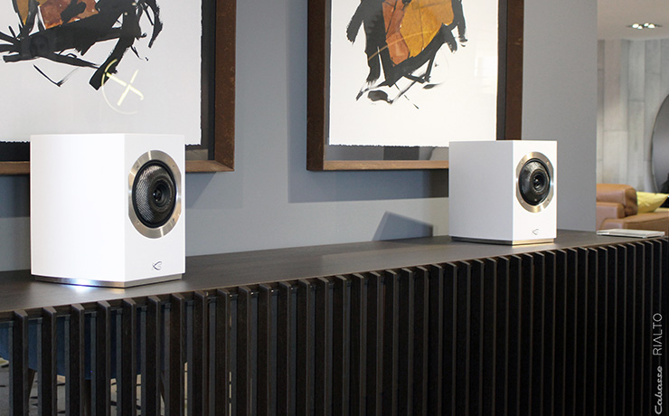 A pair of Cabasse Rialto speakers in white on a shelf with artwork behind them.