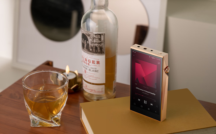 Astell & Kern A&ultima SP3000T Copper Edition Portable Music Player standing up with a bottle of something in the background and a glass full of amber coloured liquid.