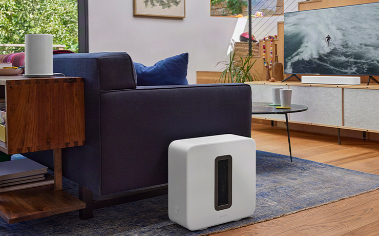 A Sonos Era 100 SL in white on a wooden unit in a living space with a Sonos Sub on the floor beside a sofa