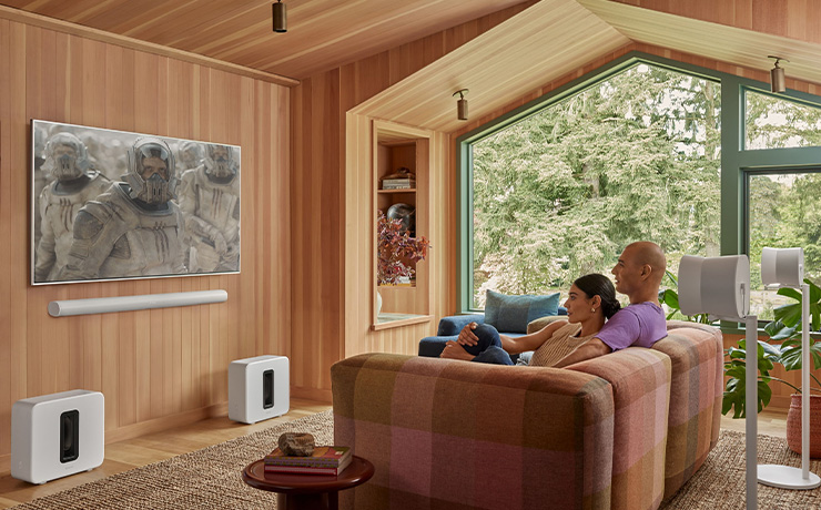 A pair of Sonos Gen 4 subs on the floor in a modern, wooden living space.  there is a man and woman sat on the sofa.  She's leaning her back on him.  there's a flat screen tv on the wall with a Sonos Arc underneath it and there are two Sonos speakers on stands behind them.  A large window looks out onto a garden.