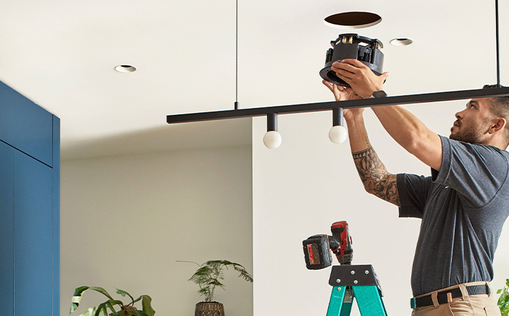 A man about to place the speaker into the cut-out hole in the ceiling. He's on a green stepladder with a drill at the top of it. He's wearing beige trousers and a dark grey top